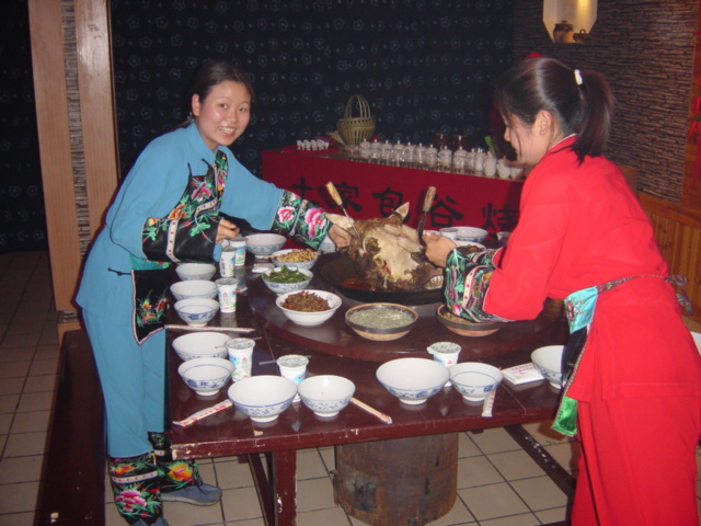 Girls dressed in traditional Tujia minority nationality dress placing a large wok containing a bull's head at the centre of the table