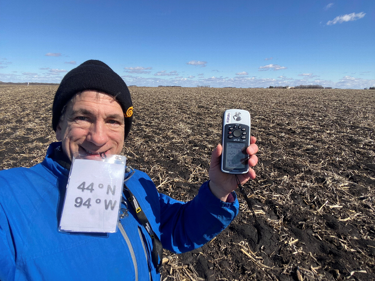 Joseph Kerski at the confluence point, holding sign in teeth due to heavy winds. 